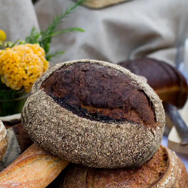 Round seeded sourdough loaves and a baguette on a linen cloth, with yellow flowers behind