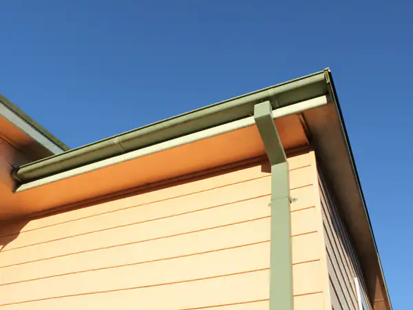 A gutter and downpipe on the corner of a house, against a blue sky.