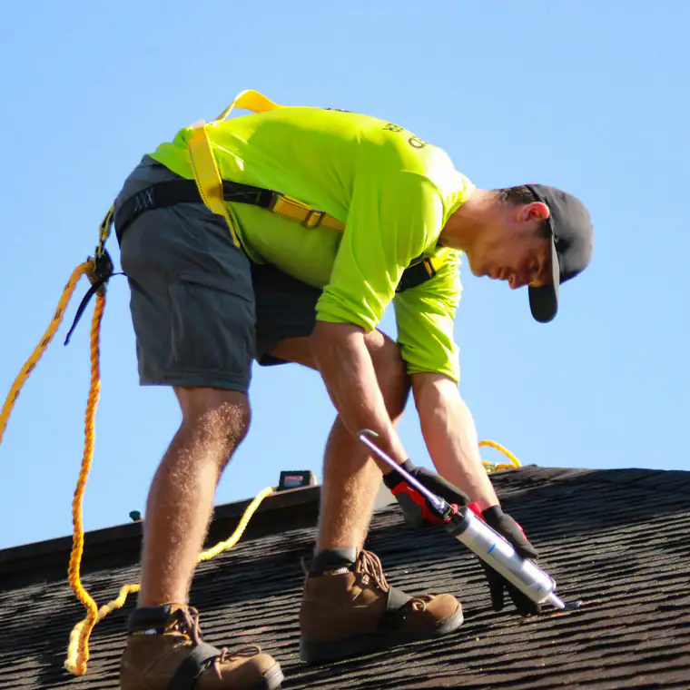 A roofer in a hi-vis shirt and safety harness seals a shingle roof under a clear blue sky