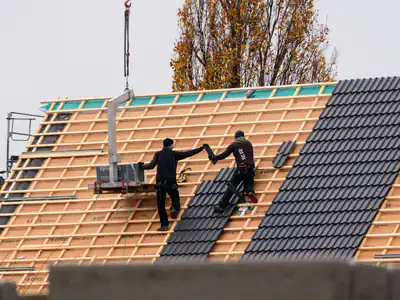 Two roofers lay dark tiles over new battens on a large pitched roof