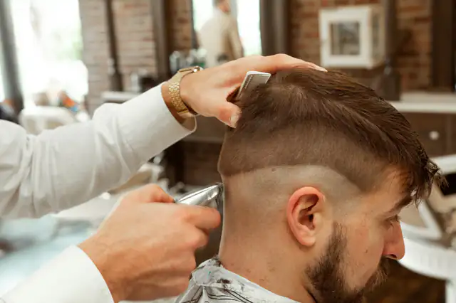 A barber trimming the side of a man’s hair into a short fade.