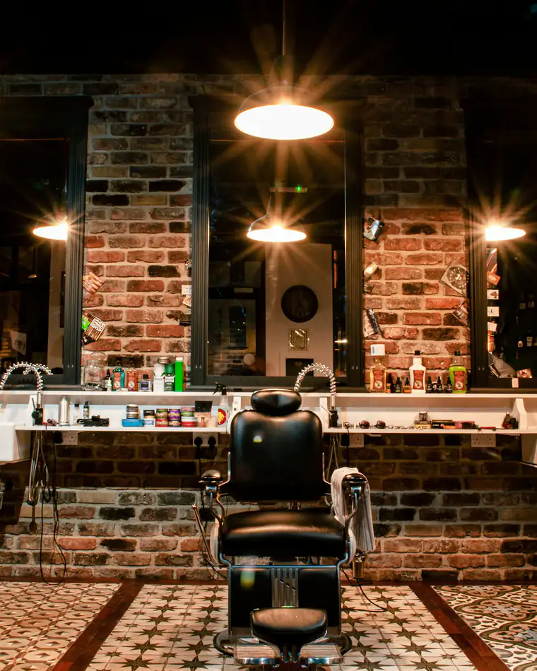 A barbershop with a black leather barber’s chair, mirrors and pendant lights against a brick wall
