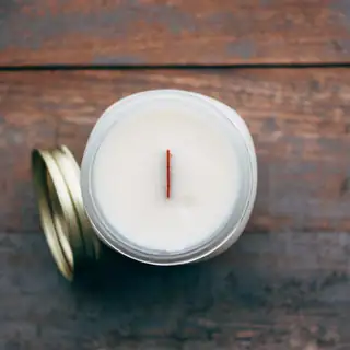 A white candle with a wooden wick in an open tin, lid beside it