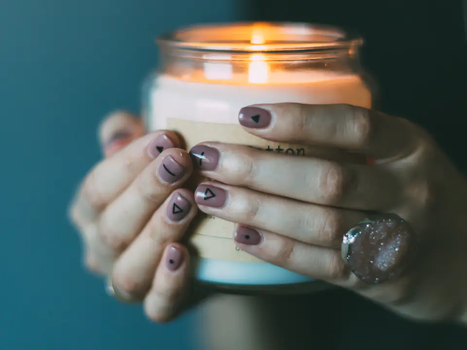 Two hands cupping a lit candle in a glass jar against a deep teal wall